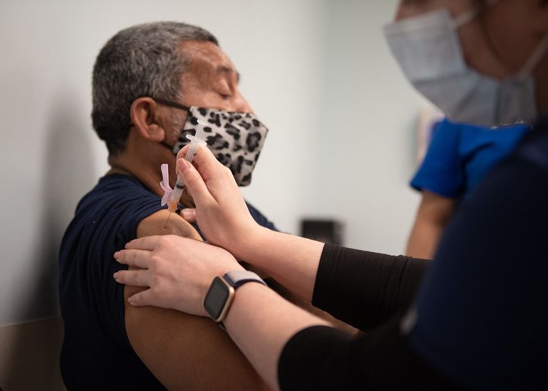 Samuel Garcia, who is seated and wears a mask, looks straight ahead as he receives a COVID-19 vaccine into his upper arm from a health care worker.