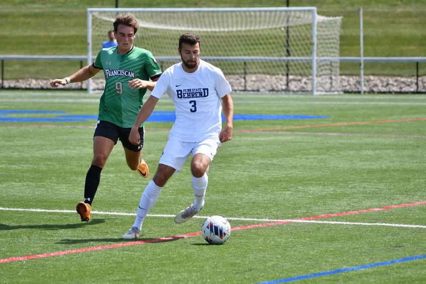 A Penn State Behrend men's soccer player advances the ball to midfield while being pressured by a defender.