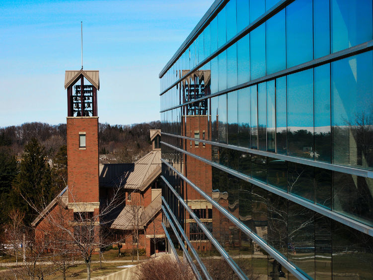 The Smith Carillon, a tall bell tower of red brick, as viewed from a building sitting beside it. The tower is reflected in the glass windows.