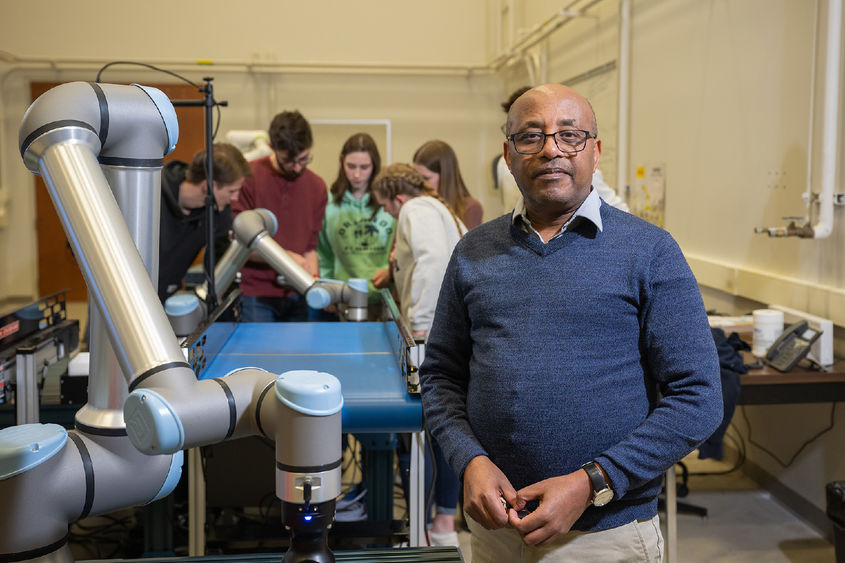 A professor stands in the Prischak Robotics and Automation Lab, with several students in the background.