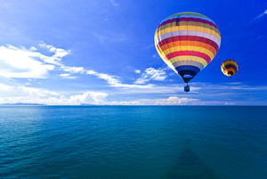 A colorful hot air balloon floats above blue water against a blue sky.