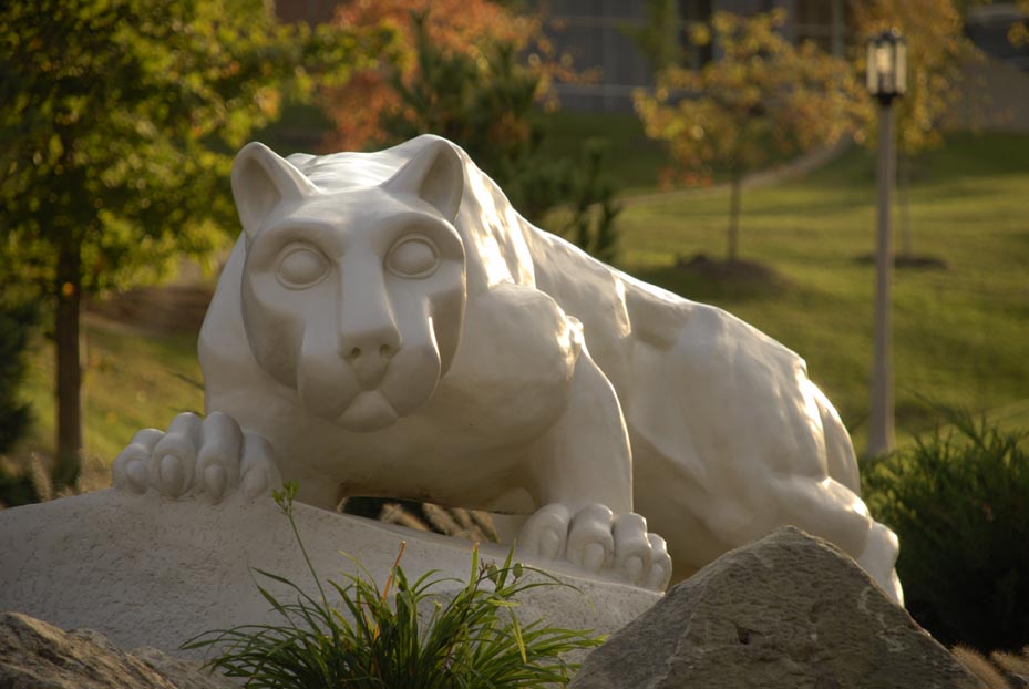 A statue of the Behrend Lion in front of autumn trees